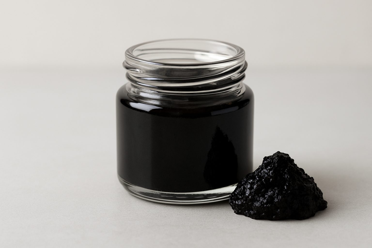 A glass jar containing dark shilajit next to a pile of the substance on a simple background